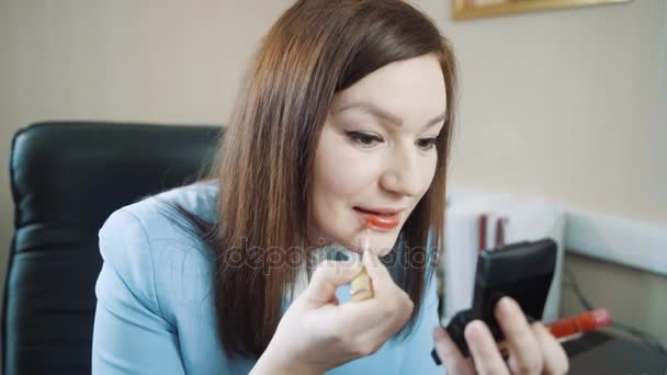 Glückliche brünette Geschäftsfrau in blauer Jacke im Büro trägt den roten Lipgloss auf. — Stockvideo