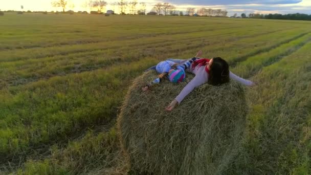 Mother with daughter lie on the straw stack on the rustic field in the sunset. — Stock Video