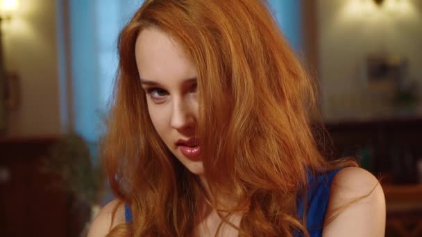 La belle fille sexy aux longs cheveux rouges et au regard ludique dans la robe bleue est assise, souriante et regardant la caméra. Slowmo . — Video