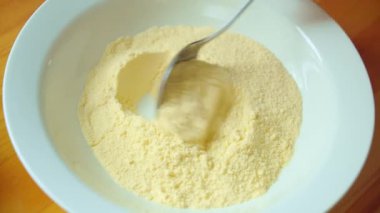 Aşçının eli ahşap masada kaşıkla beyaz kâsede sarı mısır unu karıştırır..