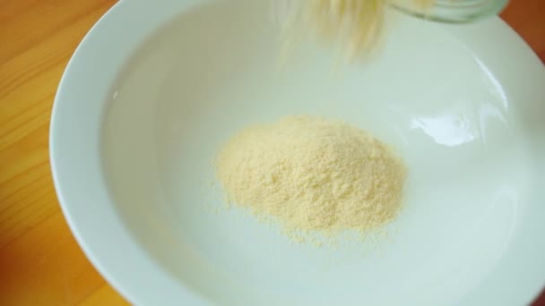 La farine de maïs jaune roule dans une assiette blanche du bocal sur une table en bois. Gros plan — Video
