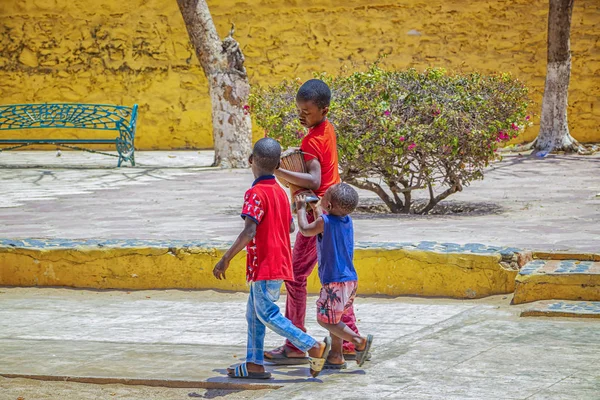 Isla Goree, Senegal 22 de abril de 2019: Niños no identificados caminan a lo largo de una calle arenosa en una pequeña ciudad y llevan un tambor de madera. — Foto de Stock