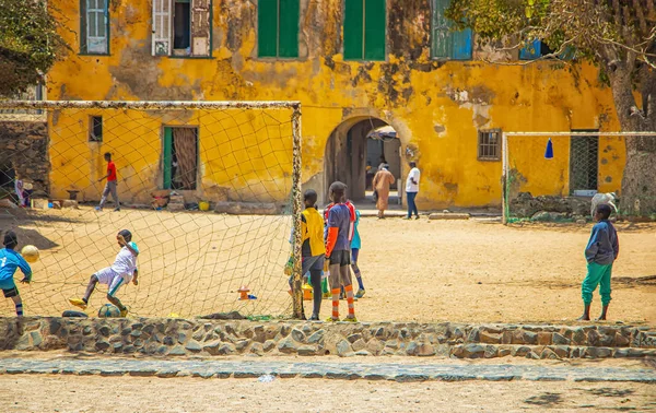 Pulau Goree, Senegal 22 April 2019: Anak laki-laki tak dikenal bermain sepak bola di pasir di kota Afrika. Bermain di depan sebuah rumah kuning tua di lapangan sepak bola. Stok Gambar Bebas Royalti