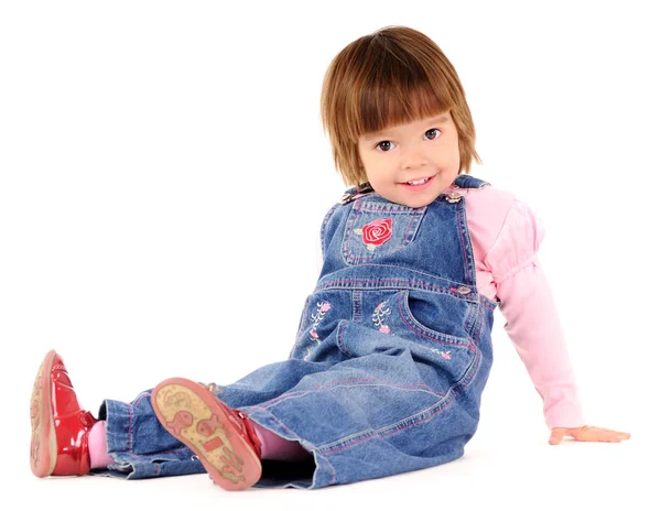 Small girl in jeans jumpsuit sitting on floor and smiling Stock Image