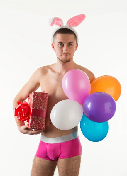 Młody mężczyzna w różowych majtkach stojący i trzymający pudełko prezentów i balony — Zdjęcie stockowe