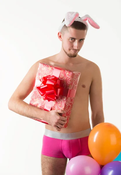 Hombre joven en ropa interior rosa para hombre de pie y sosteniendo la caja de regalo y globos — Foto de Stock