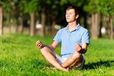 Genç adam parkta yeşil çimlerde oturuyor ve meditasyon yapıyor.