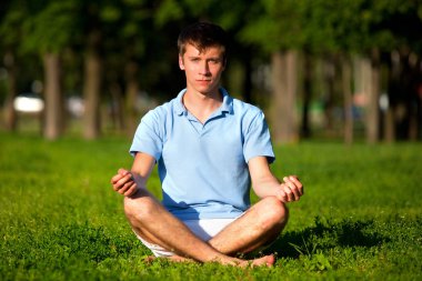 Genç adam parkta yeşil çimlerde oturuyor ve meditasyon yapıyor.