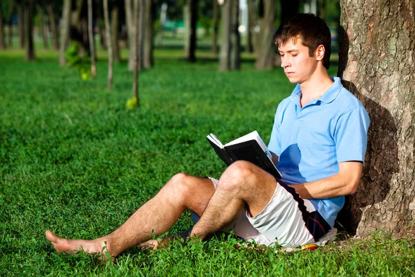 Giovane seduto sull'erba verde vicino all'albero e libro di lettura — Foto Stock