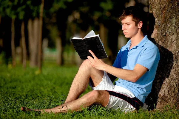 Młody człowiek siedzi na zielonej trawie obok drzewa i czyta książkę — Zdjęcie stockowe