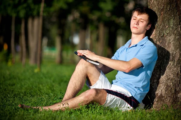 Joven sentado sobre hierba verde cerca del árbol y leyendo libro —  Fotos de Stock