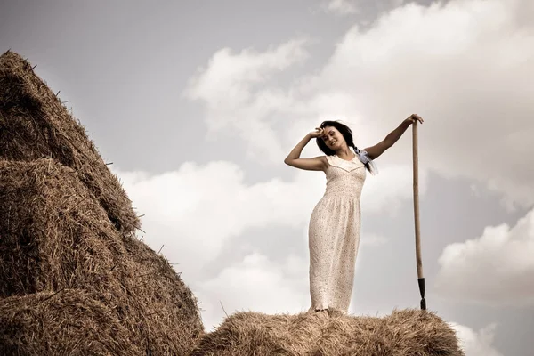 Молодая женщина в белом платье стоит с вилами сена в руке в поле — стоковое фото