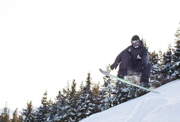 Giovane uomo in abbigliamento sportivo snowboard su sfondo pini — Foto Stock