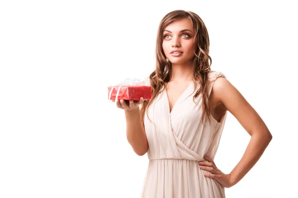 Young beautiful positive brunette woman in beige dress standing, holding small red gift box in hand and smiling — Stock Photo, Image