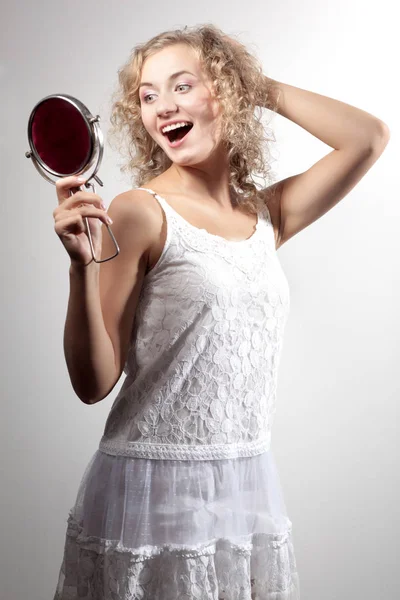 Young slim beautiful blond woman with curly hair in white sexy mini dress looking at small mirror and smiling — Stock Photo, Image