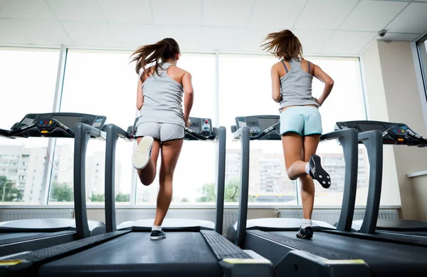 Dos jóvenes hermosas mujeres delgadas en ropa deportiva corriendo en las cintas de correr en el gimnasio con ventanas y la ciudad en el fondo —  Fotos de Stock