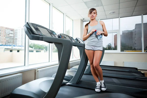 Young beautiful slim women in grey sportswear standing on treadmill with bottle of water in gym — Stock Photo, Image