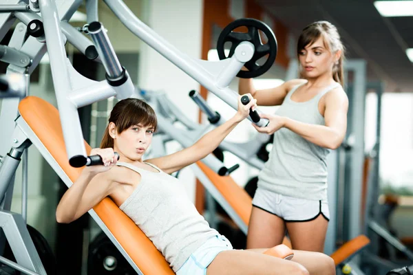 Jovem bela mulher morena instrutora em sportswear ajudando outra menina com o uso de máquina de fitness para braços no ginásio — Fotografia de Stock