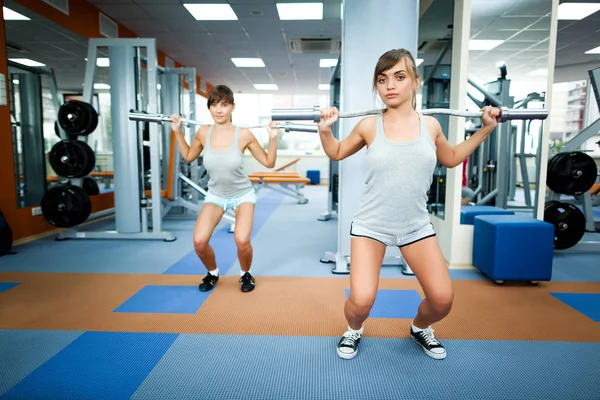 Jeune femme brune souriante instructrice en vêtements de sport gris montrant comment faire squat d'haltère à une autre fille dans la salle de gym — Photo