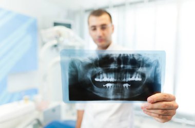 Beyaz üniformalı genç bir dişçi klinikteki dişçi muayenehanesinde durup diş fotoğrafına bakıyor.