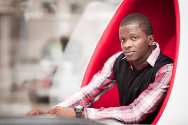 Büyük kırmızı yumurta şekilli koltukta oturan şık giysili Afrikalı genç adam.
