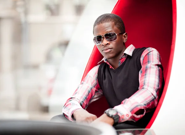 Joven hombre africano guapo en ropa casual elegante y gafas de sol sentado en un gran sillón en forma de huevo rojo —  Fotos de Stock