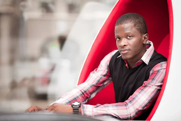 Joven hombre africano guapo en ropa casual elegante sentado en un gran sillón en forma de huevo rojo —  Fotos de Stock