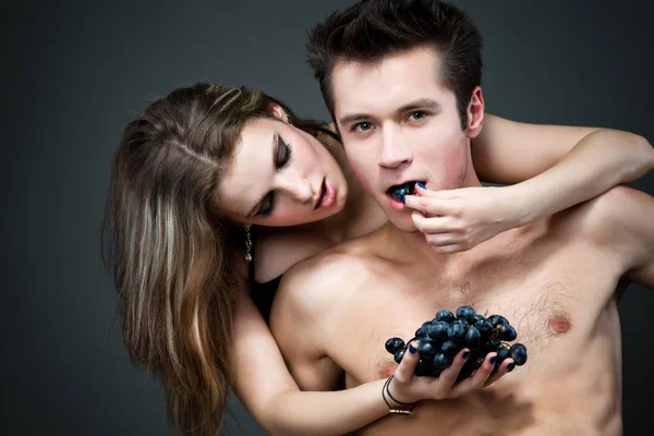 Genç, gülümseyen komik adam arka planda çıplak kız arkadaşıyla siyah üzüm yiyor koyu gri arka planda. — Stok fotoğraf