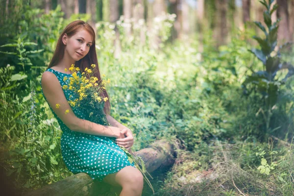 Nő zöld ruhában ül a rönk és a gazdaság sárga virágok az erdőben — Stock Fotó