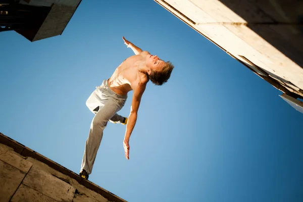 Joven saltando y practicando parkour entre edificios en el exterior en un día claro de verano con el cielo azul al fondo —  Fotos de Stock