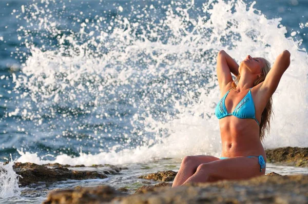 Young blond woman in blue bikini sitting on rocks and enjoying drops of waves — 스톡 사진