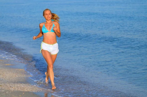 Młoda kobieta w odzieży sportowej biegnie w pobliżu brzegu morza i uśmiecha się — Zdjęcie stockowe