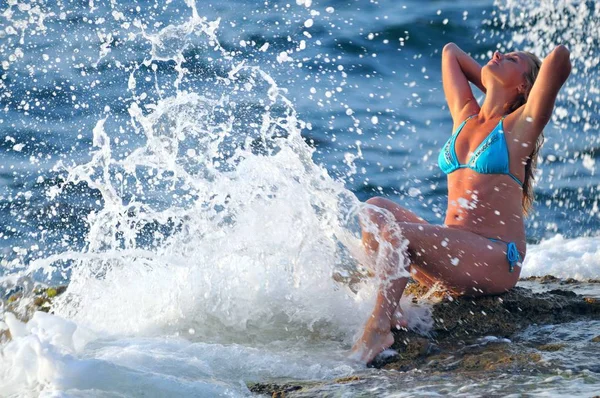 Junge blonde Frau im blauen Bikini sitzt auf Felsen und genießt Tropfen von Wellen — Stockfoto