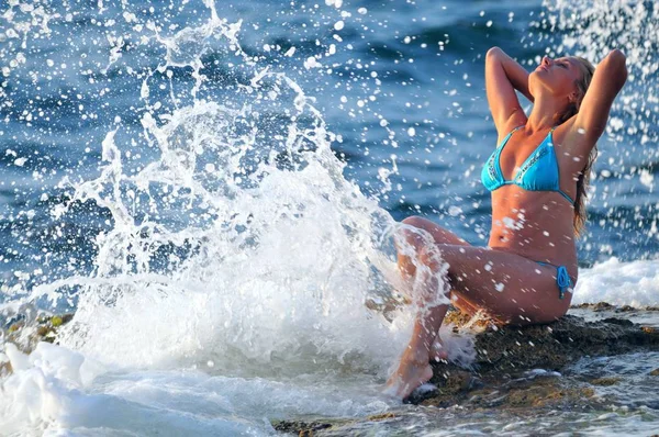 Mladá blond žena v modrých bikinách sedí na skalách a těší kapky vln — Stock fotografie