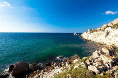 Durgun mavi deniz suları kıyıları, kayalık kıyı şeridi ve parlak güneş.