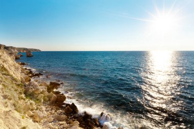 Durgun mavi deniz suları kıyıları, kayalık kıyı şeridi ve parlak güneş.