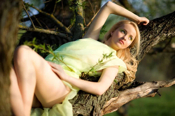Young sexy woman in green dress sitting on tree trunk over water on summer day with green nature at background — 스톡 사진
