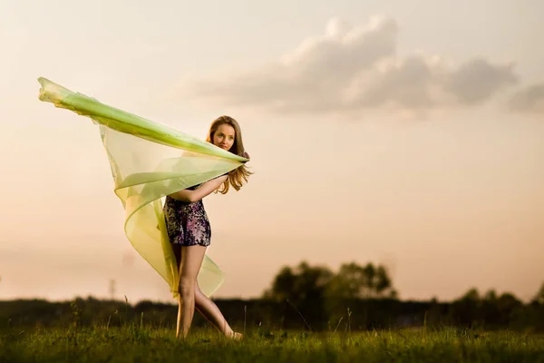 Mujer joven en vestido haciendo pose gimnástica y sosteniendo tela amarilla en el día de verano con paisaje de campo en el fondo — Foto de Stock