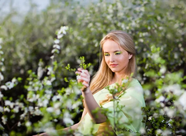 Young beautiful blond smiling woman in yellow dress standing in blooming cherry trees and looking at flower — 스톡 사진