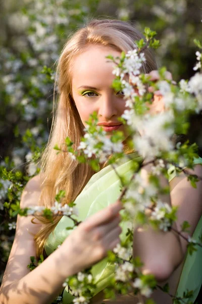 Mladá krásná blond usmívající se žena ve žlutých šatech stojící v kvetoucích třešních stromů a při pohledu na květinu — Stock fotografie