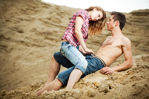 Young woman in jeans sitting on her boyfriend man and unfastening his belt on sandy ground on summer day — 스톡 사진