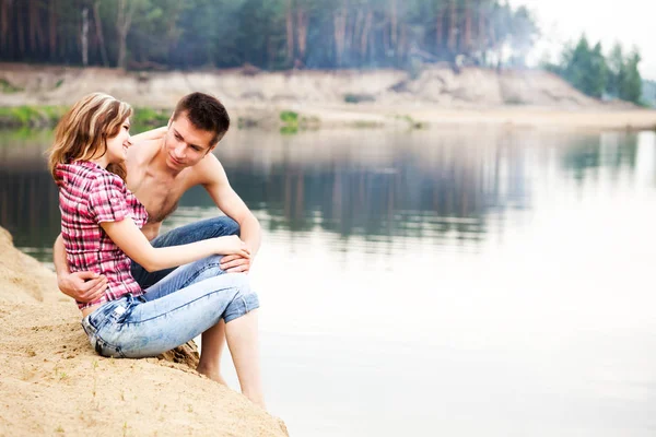 Jong mooi liefdevol paar met rust op de oever van de rivier op zomerse dag met groene natuur op de achtergrond — Stockfoto