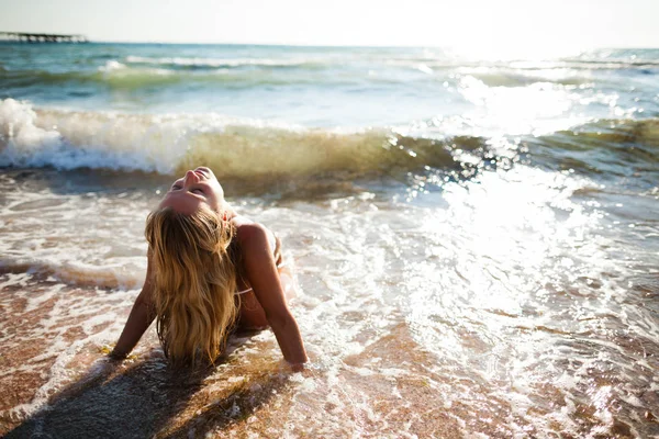 Молода красива блондинка в білому бікіні сидить на краю морської води в хвилях піни і насолоджується сонячним сяйвом — стокове фото
