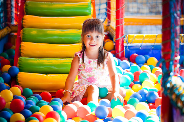 Small smiling girl in dress sitting in colorful soft decorative balls in playroom