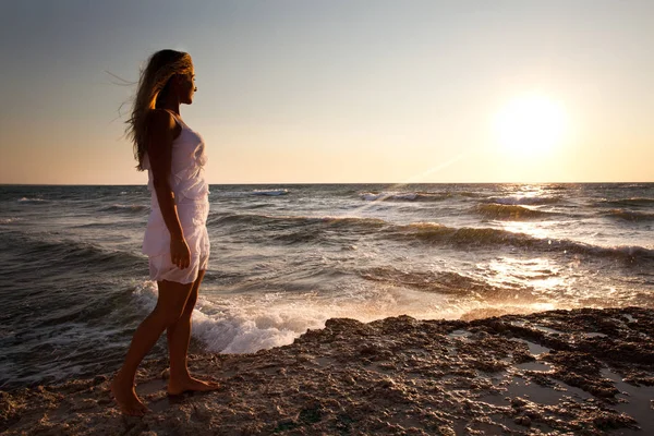 Jovem mulher loira sorridente em vestido branco em pé sobre rochas e olhando para o pôr do sol sobre a água do mar ondulada — Fotografia de Stock