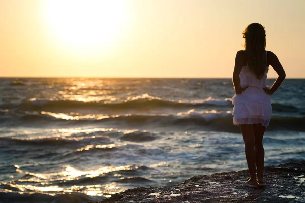 Young smiling blond woman in white dress standing on rocks and looking at sunset over wavy sea water — Stock Photo, Image