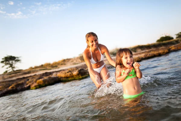 Joven mujer rubia en bikini blanco y pequeña chica feliz divirtiéndose en agua de mar con playa rocosa en el fondo — Foto de Stock