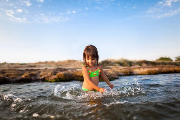 Pequeña chica sonriente en traje de baño brillante de pie y jugando con agua de mar con fondo de playa rocosa —  Fotos de Stock