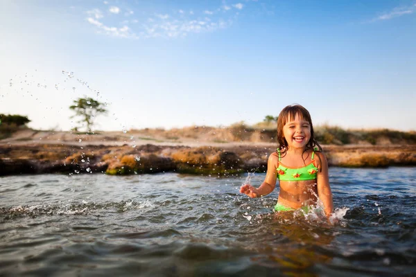 Pequeña chica sonriente en traje de baño brillante de pie y jugando con agua de mar con fondo de playa rocosa — Foto de Stock