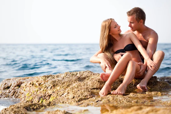 Jong gelukkig mooi paar vrouw en man zitten op rotsen en kijken naar elkaar met zee op de achtergrond — Stockfoto